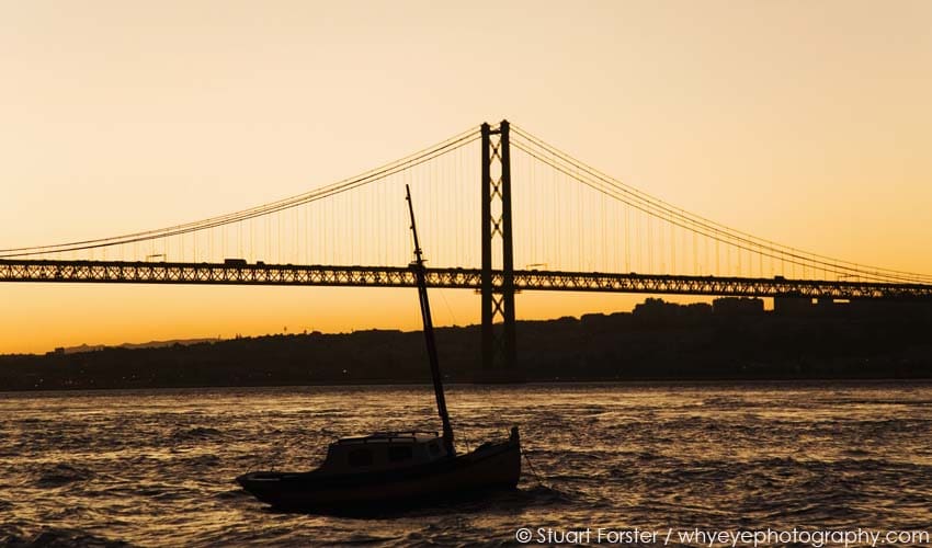 Boat on the River Tagus, Rio Tejo, silhouetted during golden dusk under 25 April Bridge, Ponte de 25 Abril, Lisbon, Portugal