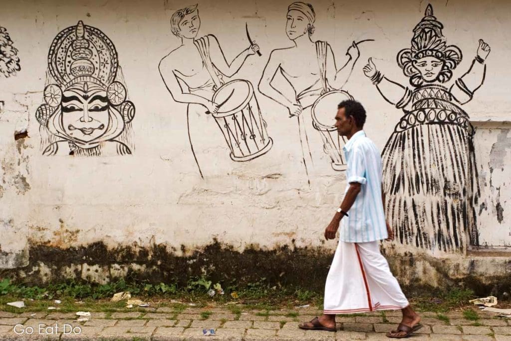 A man wearing a dhoti walking past a wall with symbols of traditional heritage in Kerala.