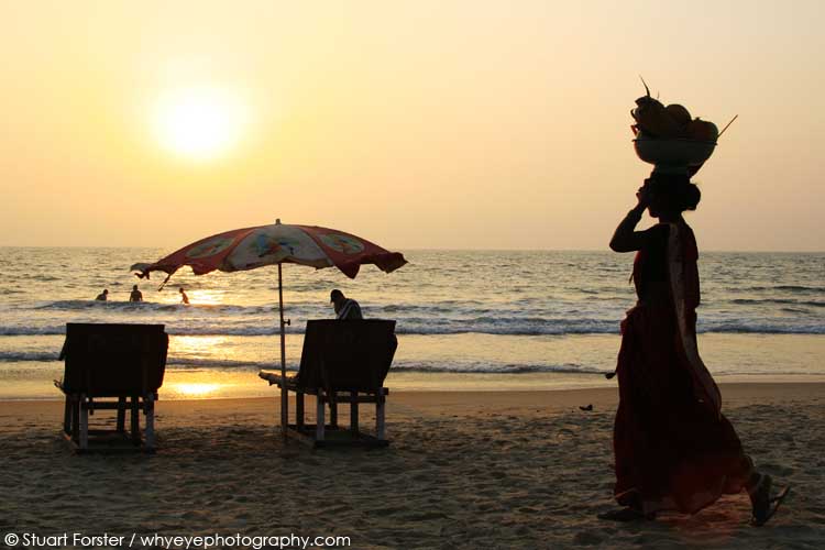 At sunset a silhouetted woman carries a basket on her head at the sun sets over Bogmalo Beach in Goa, India