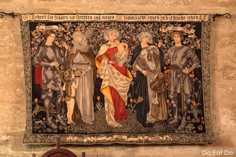 Tapestry depicting figures in medieval costumes with German text on a stone wall at Langley Castle in Northumberland, one of the castle hotels in England