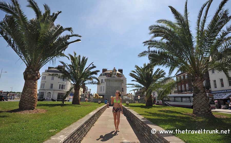 Woman walking between palm trees on a sunny day at the Regency seafront in Weymouth, Dorset