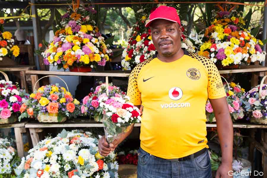 Smiling florist wearing a Kaizer Chiefs football shirt at a flower stall at African Unity Square in Harare, Zimbabwe