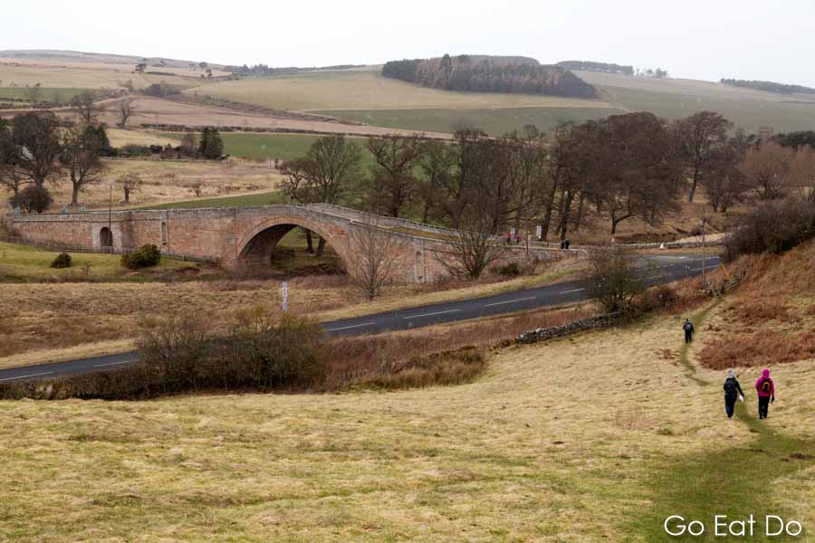 Walkers walking towards Weetwood Bridge, a stone humpback structure, in the Cheviot Hills of Northumberland in northeast England