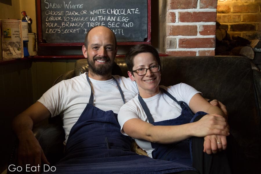 Chefs Guy and Brittany Manning smiling while sitting on the fireside sofa at their pub and restaurant.