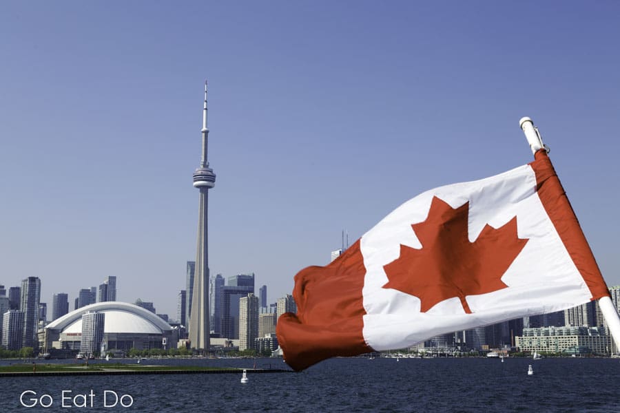 A Canadian maple leaf flag flutters in front of the CN Tower on a sunny day in Toronto, Ontario
