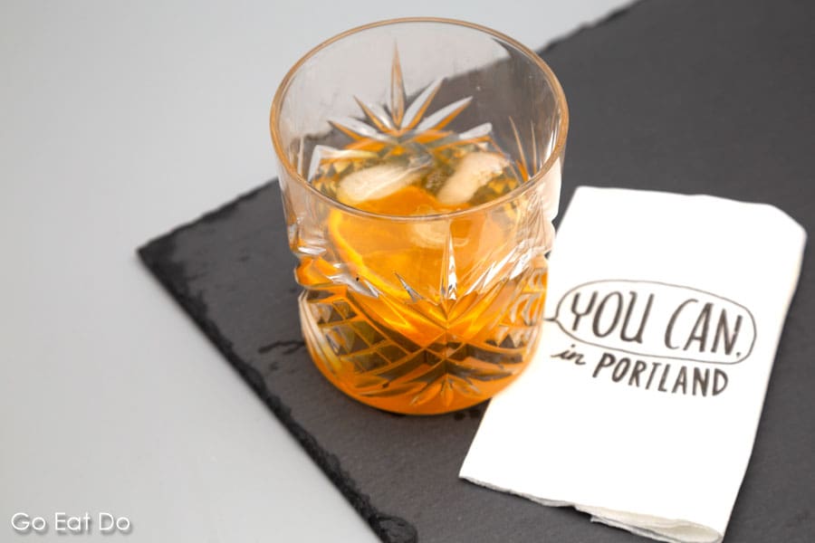 An Old Fashioned cocktail, made with whiskey, garnished with a slice of orange on a slate next to a 'You can in Portland' napki