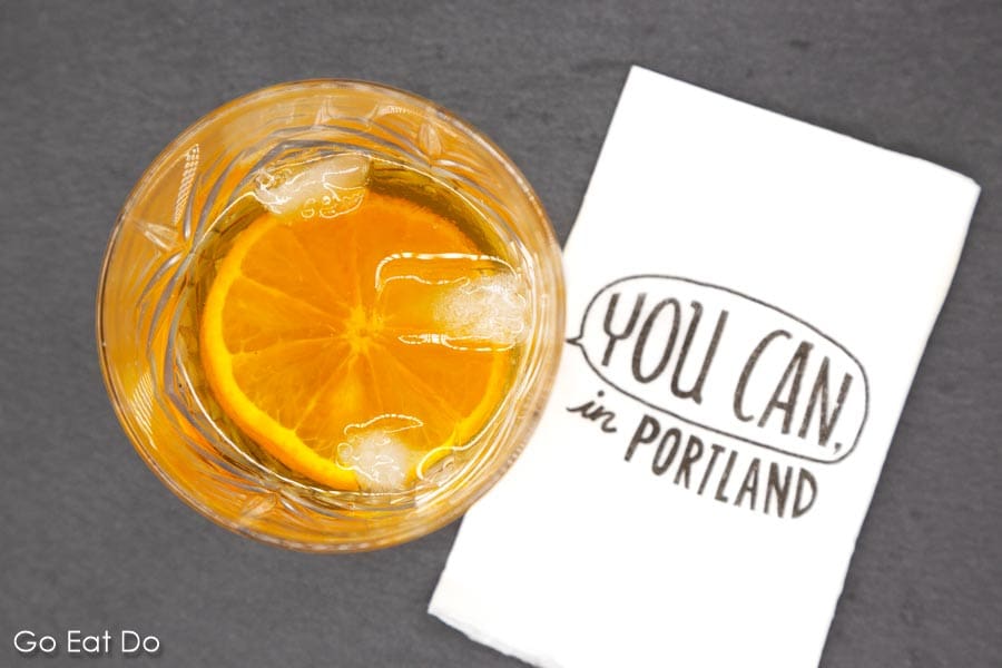 A whisky cocktail garnished with orange on a slate next to a 'You can in Portland' napkin