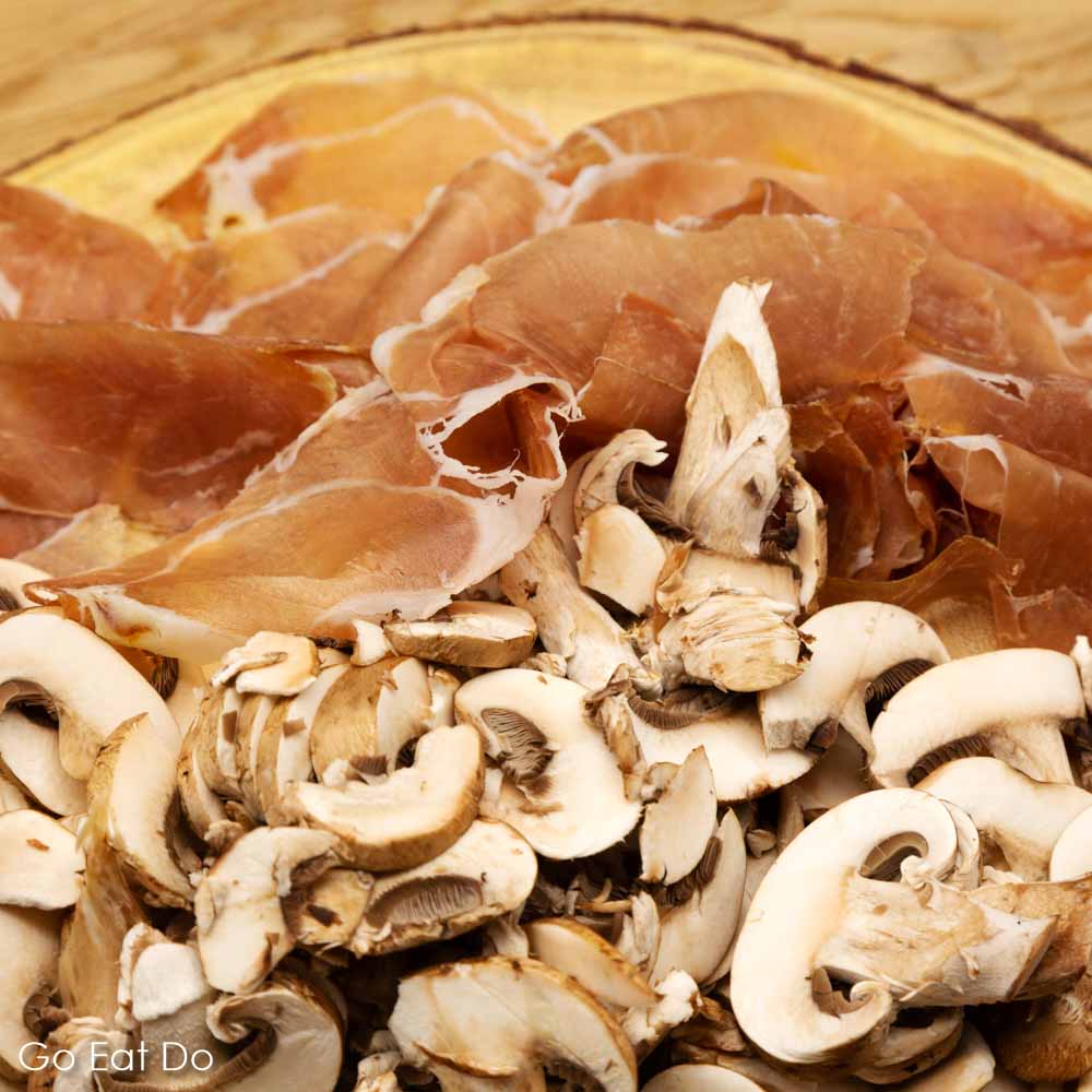 Slices of Parma ham (Prosciutto di Parma) and slice brown mushrooms presented on a wooden board