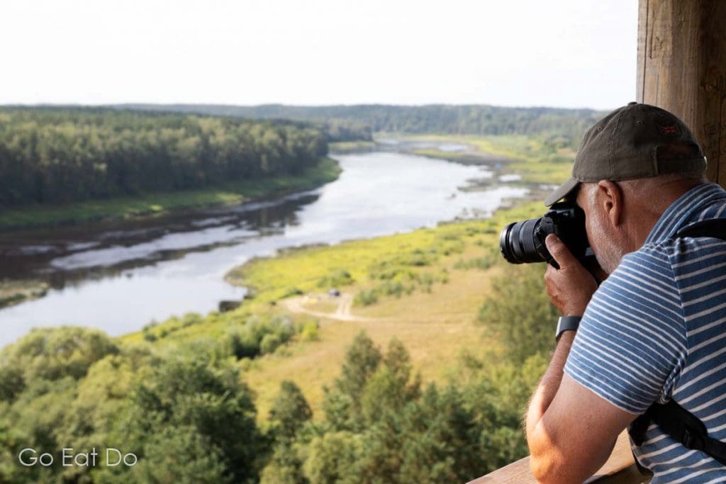 Man photographs the landscape of the Daugavas Loki Nature Park from Vasargelišķi tower whose viewing platform overlooks the Daugava River meandering through the Daugava Valley.