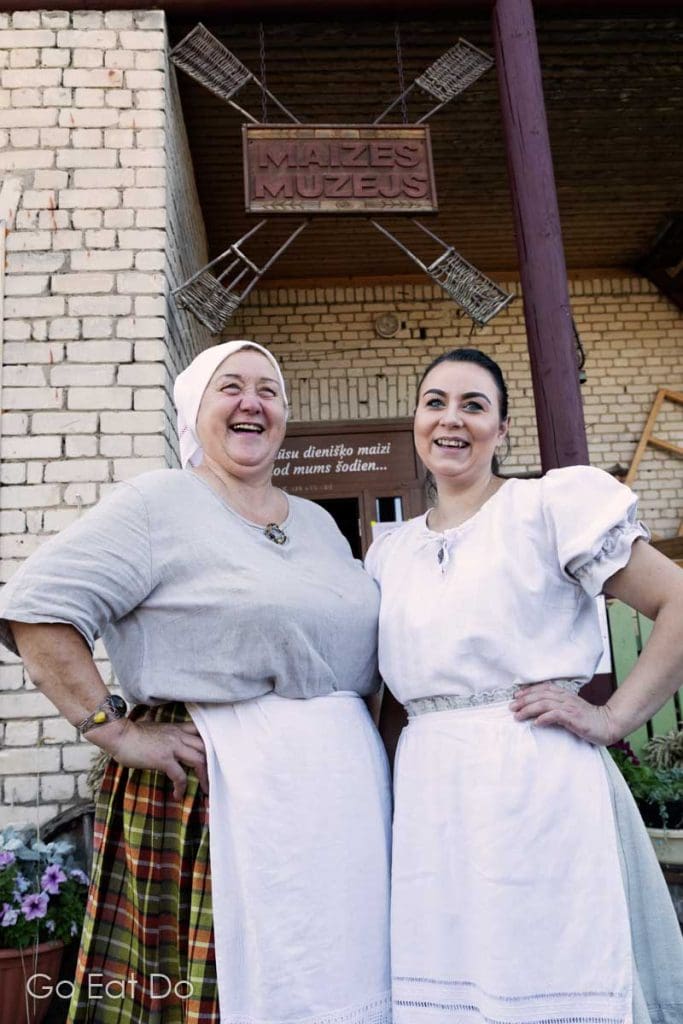 Women in traditional Latgalian costumes smiling and welcoming guests to the Bread Museum in Aglona, Latvia.