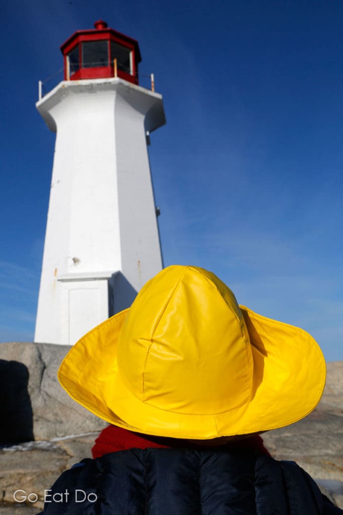 A person wearing a yellow souwester hat looks at Peggy's Cove Lighthouse on a sunny day.