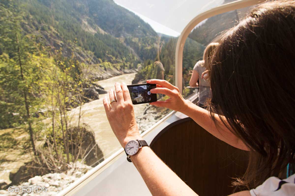 Woman photographs the landscape of British Columbia through the panoramic window on the upper level of a Rocky Mountaineer GoldLeaf Service car.